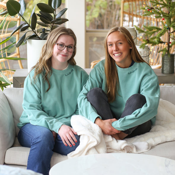 Two models wearing aqua long sleeve crewneck sweatshirt with embroidered "Homebody" around the collar. 