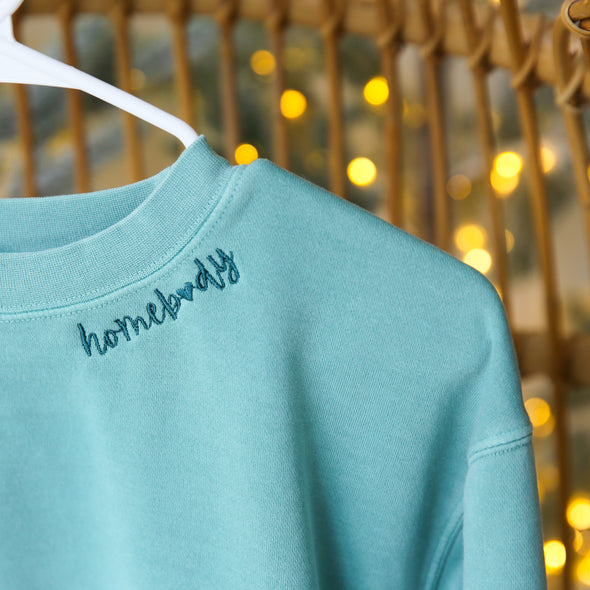 Aqua long sleeve crewneck sweatshirt with embroidered "Homebody" around the collar. 